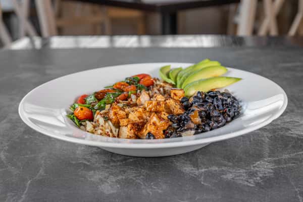 Breakfast bowl featuring seasoned grains, black beans, diced vegetables, fresh herbs, and sliced avocado, served on a white plate.