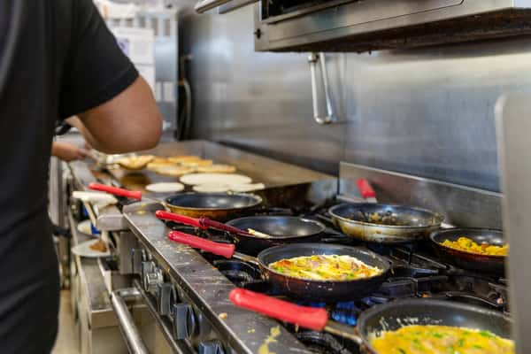 Restaurant chef preparing multiple dishes on a busy commercial kitchen stovetop, with frying pans, cooking utensils, and stainless steel equipment.