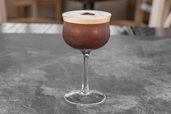 Chocolate-colored cocktail served in a stemmed glass with a thick layer of creamy foam and a dusting of cocoa powder, placed on a dark tabletop.