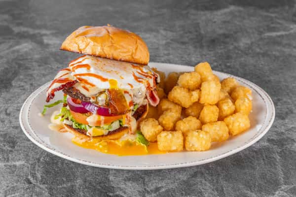 Juicy cheeseburger stacked with lettuce, tomato, red onion, melted cheese, and a toasted bun, served with crispy golden tater tots on a white plate.