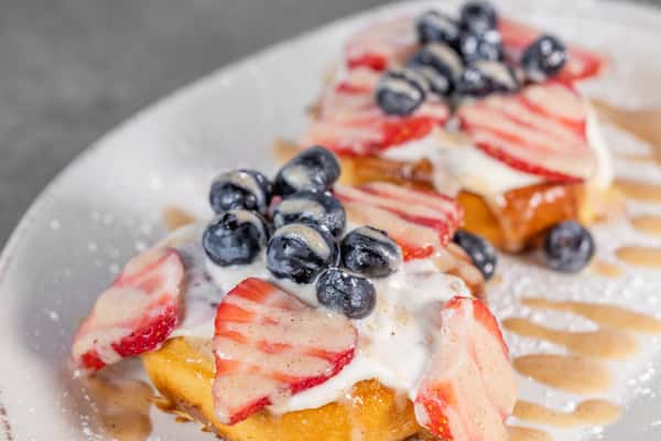 Close-up of golden French toast topped with sliced strawberries, fresh blueberries, and a generous drizzle of creamy white sauce.