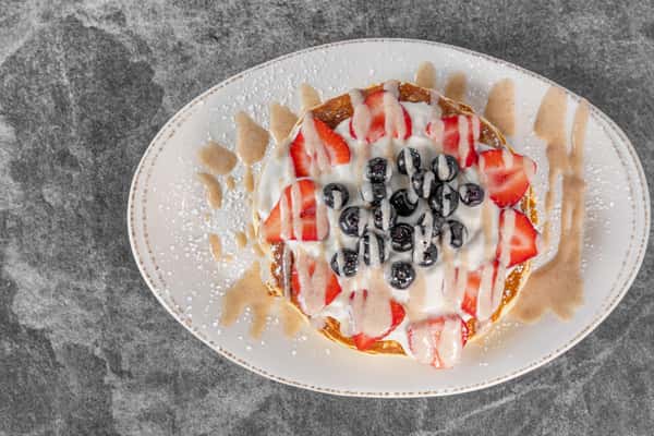 Overhead view of French toast topped with sliced strawberries, blueberries, whipped cream, and a dusting of powdered sugar on a white plate.