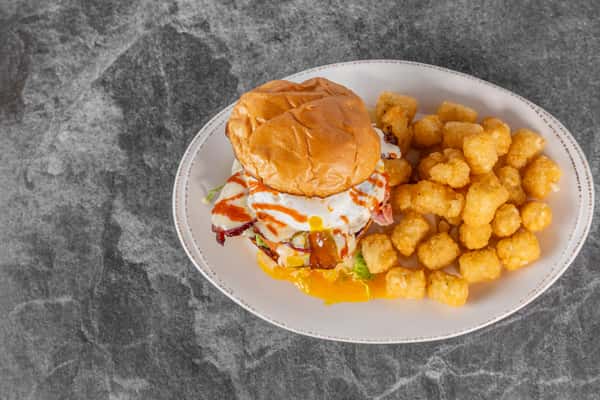 Breakfast sandwich filled with eggs, melted cheese, and savory toppings on a toasted bun, served with crispy golden tater tots on a white plate.