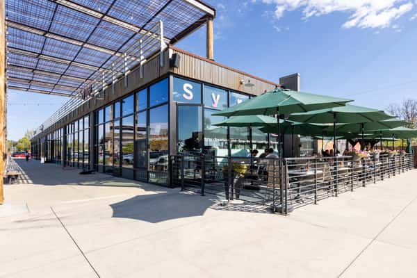 Exterior view of a restaurant with a spacious outdoor dining patio, black tables and chairs, large green umbrellas, and a metal staircase under a clear blue sky.