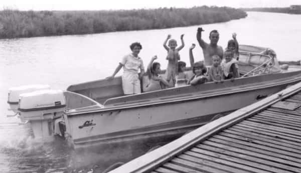 Vintage black-and-white photo of a family riding in a small motorboat on a Louisiana bayou, waving and smiling near a wooden dock, capturing nostalgic coastal dining and fishing culture