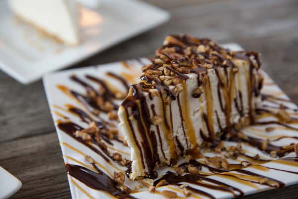 Slice of creamy cheesecake topped with chopped nuts, drizzled with chocolate and caramel sauce on a white plate, rustic restaurant dessert presentation