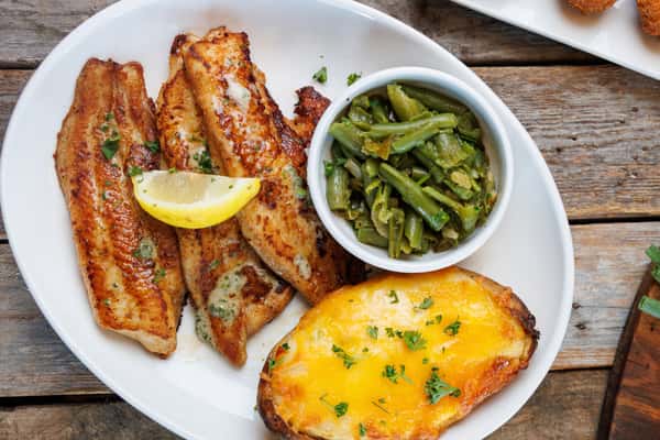 Blackened fish fillets seasoned with Cajun spices, served with sautéed green beans, a cheesy baked potato, and lemon garnish on a casual Southern-style plate