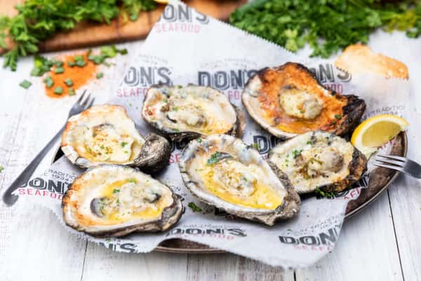 Grilled oysters topped with garlic butter, herbs, and melted cheese, served on the half shell with lemon wedges on a rustic seafood restaurant platter