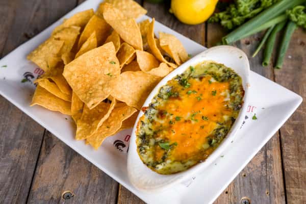 Warm spinach artichoke dip topped with melted cheddar cheese, served with crispy tortilla chips on a rustic wooden table