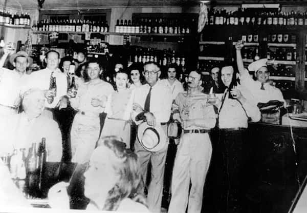 Historic black-and-white interior scene of a lively bar at Don's Seafood, with patrons raising drinks and shelves of bottles in the background, showcasing classic Cajun dining atmosphere