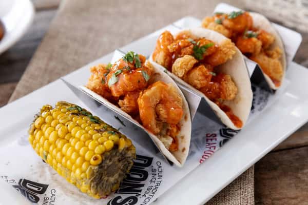 Crispy shrimp tacos drizzled with spicy orange sauce and topped with fresh herbs, served in soft tortillas alongside grilled corn on the cob on a casual seafood restaurant plate