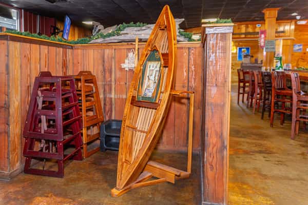 This image showcases the same wooden canoe-themed decorative piece with fishing gear artwork seen earlier, now shown in its full setting. It's placed beside a stack of high chairs in a rustic-style restaurant interior, with visible booths, bar stools, and a well-lit wooden decor theme.