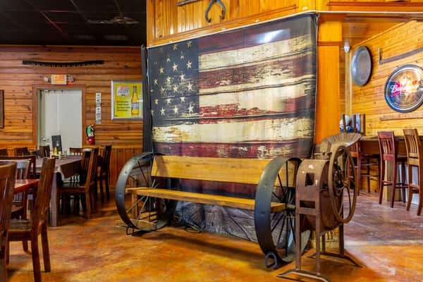 Wagon wheel bench with wooden seat in front of a vintage American flag backdrop inside a rustic wood-paneled restaurant.