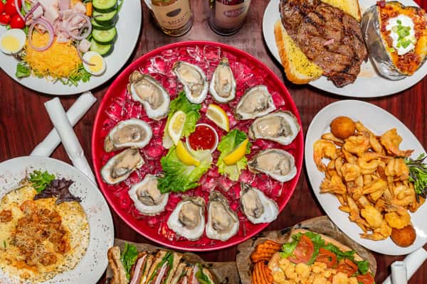 Seafood platter with raw oysters on ice, lemon wedges, cocktail sauce, and assorted dishes like steak, shrimp, salad, and pasta.