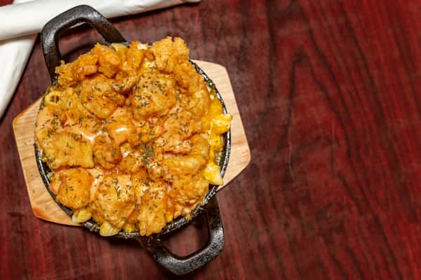 Skillet of creamy macaroni and cheese topped with crispy fried seafood and drizzled with sauce on a wooden serving board.