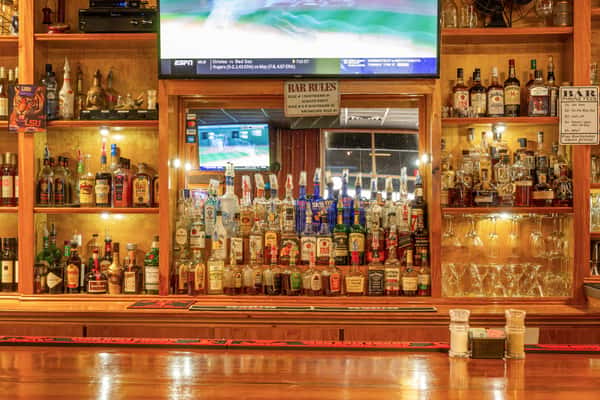 Well-stocked wooden bar with liquor bottles, glassware, and a TV showing a baseball game, viewed from across the polished counter.