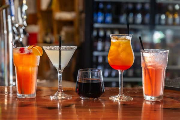 Variety of colorful cocktails and beverages lined up on a wooden bar counter with a blurred background of a bar setting.