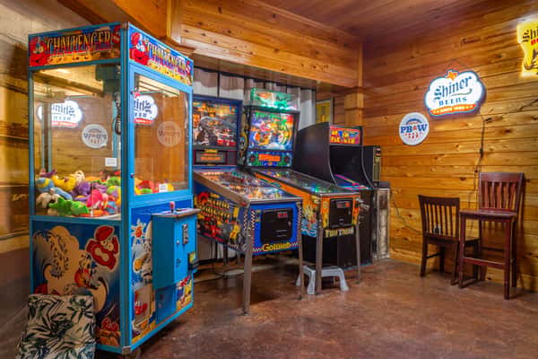 Corner arcade with a claw machine, two pinball machines, and a classic Ms. Pac-Man game, surrounded by wood-paneled walls and bar signs.