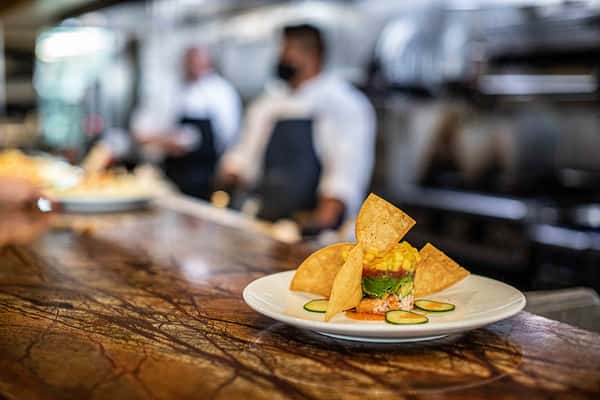 A carefully plated appetizer sits on a wooden bar, featuring crisp, golden elements arranged with precision. In the softly blurred background, restaurant staff move through a busy kitchen, adding a sense of energy and craftsmanship to the scene.