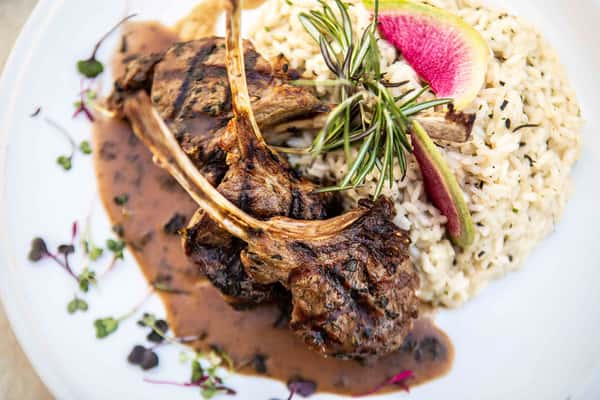 A beautifully seared rack of lamb is the centerpiece of this elegant dish, with the bones standing upright for a dramatic presentation. The meat is perfectly charred on the outside and garnished with fresh rosemary, adding a rustic, aromatic touch. It's served alongside creamy mashed potatoes and a vibrant vegetable garnish, all finished with a rich sauce drizzled over the plate. The composition and detail suggest a high-end, gourmet dining experience.