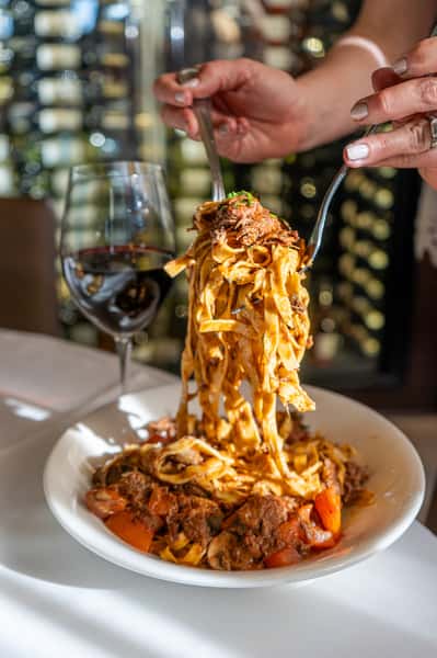 A rich and indulgent pasta dish is being lifted from the plate, with strands coated in a hearty meat sauce and topped with melted cheese. The motion captures the stretch and texture of the pasta, while a glass of red wine in the background enhances the warm, inviting dining experience.