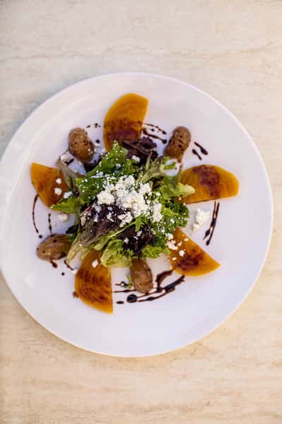 A beautifully plated gourmet dish is presented on a white plate, featuring vibrant orange elements arranged with precision. It's garnished with fresh herbs and crumbled cheese, with a rich balsamic drizzle adding contrast and elegance—highlighting a refined, artistic approach to Italian cuisine.