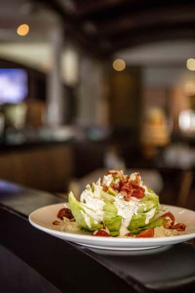 A fresh, colorful salad is plated with crisp greens, sliced vegetables, and a generous topping of cheese. The softly blurred restaurant background enhances the focus on the dish, emphasizing freshness and simplicity in a relaxed dining environment.