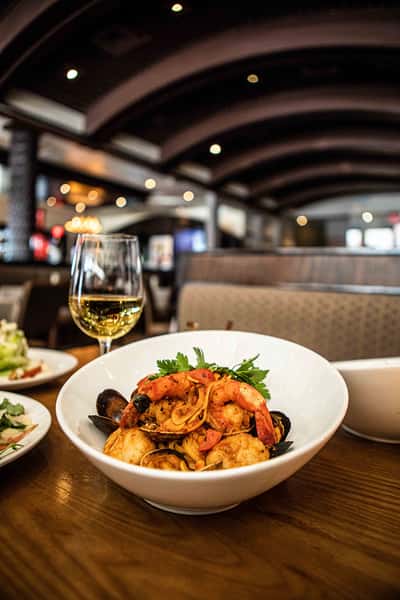 A bowl of pasta sits on a restaurant table alongside a glass of white wine. The setting features a modern, warmly lit dining room with curved architectural details, creating a comfortable yet upscale atmosphere perfect for enjoying a flavorful meal.