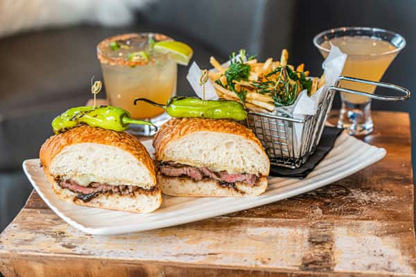 Grilled steak sandwich topped with melted cheese and roasted pepper on artisan bread, paired with truffle fries and signature cocktails — upscale gastropub presentation.