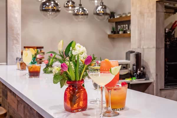 Colorful cocktails garnished with fruit sit on a modern white bar, decorated with fresh tulips and ambient pendant lighting.
