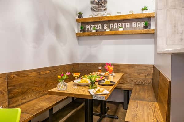 Cozy corner booth with wooden benches, colorful drinks, and Italian dishes beneath decorative shelves labeled Pizza & Pasta Bar.