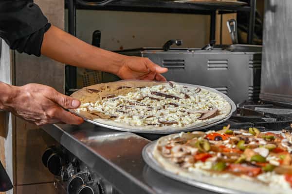 Chef placing an uncooked mushroom and cheese pizza into an oven, with a colorful veggie pizza in the foreground.