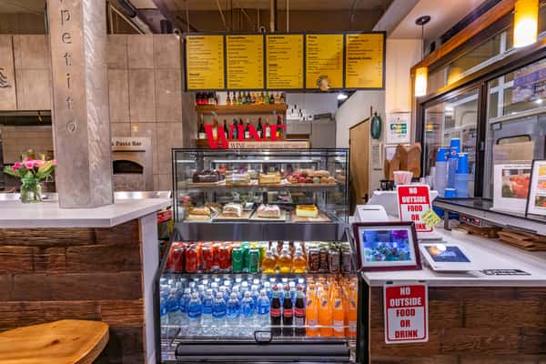 Dessert and drink display with cakes, pastries, bottled beverages, and a visible overhead menu at a pizza and pasta bar counter.