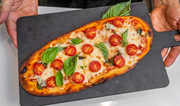 Flatbread pizza topped with melted mozzarella, cherry tomatoes, and fresh basil leaves on a black serving board.