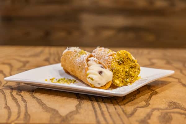 Two cannoli filled with creamy ricotta filling, one end dipped in crushed pistachios and lightly dusted with powdered sugar, served on a white rectangular plate.