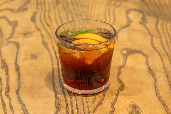 Iced dark beverage in a glass, garnished with orange slices and mint, served on a wood-patterned table.