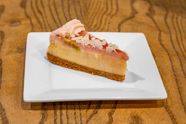 Slice of cheesecake with pink frosting, white chocolate shavings, and graham cracker crust on a square white plate.