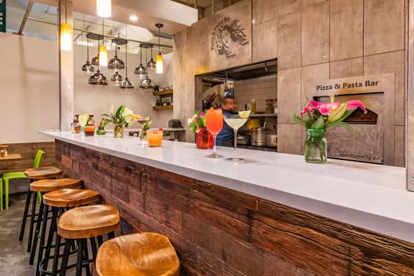 Bar counter with wood paneling, colorful cocktails, fresh flowers, pendant lights, and a visible kitchen at a pizza and pasta bar.