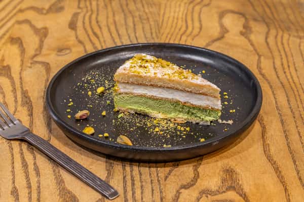 Slice of layered pistachio cake topped with crushed nuts, served on a black plate with scattered pistachio pieces.