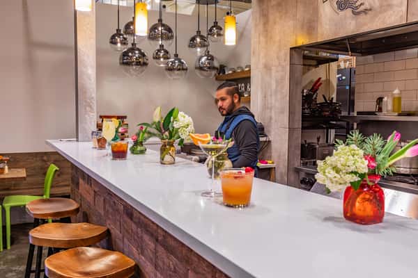 Modern bar with wooden stools, colorful cocktails, floral decor, and a bartender behind the counter near an open kitchen.