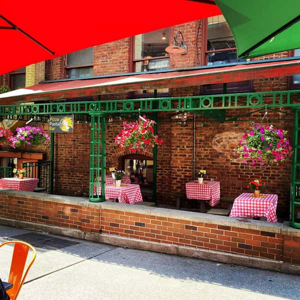 Outdoor patio with red-checkered tables, vibrant hanging flower baskets, and green metal framing outside a brick Pizza & Pasta Bar.