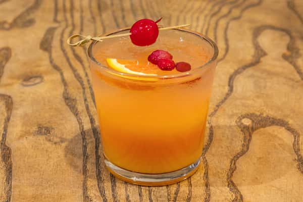 Orange cocktail garnished with a cherry and orange slice, served over ice in a short glass on a wooden table.
