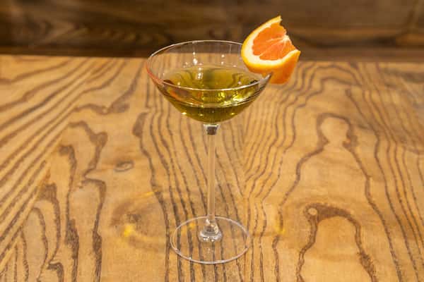 Martini glass with a green cocktail, garnished with an orange slice, on a wooden table.