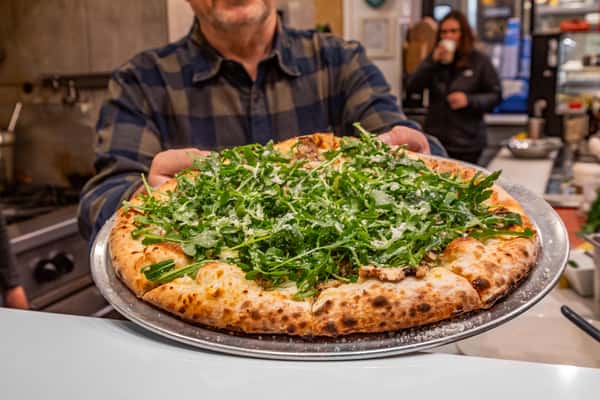 Wood-fired pizza topped with fresh arugula and grated cheese on a metal tray, held by a person in a plaid shirt.