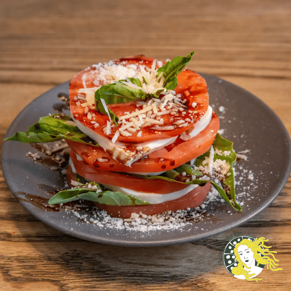 Stacked Caprese salad with fresh tomato slices, mozzarella, basil leaves, balsamic glaze, and grated cheese on a dark plate.