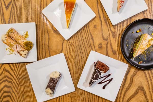 Assorted Italian desserts on plates, including cannoli, pistachio cake, flan, and Oreo chocolate cake on a wooden table.