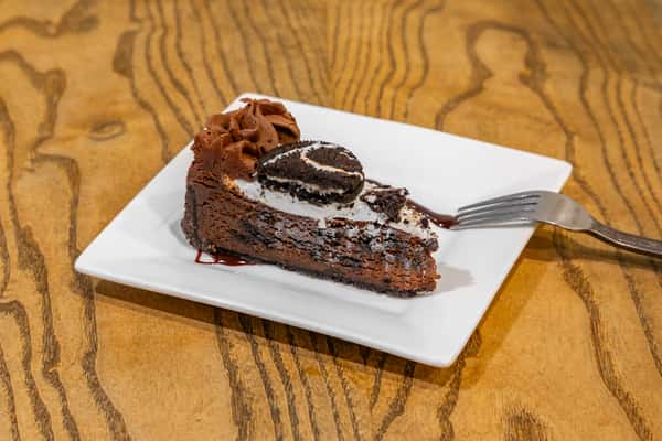 Slice of rich chocolate Oreo cake topped with whipped cream, chocolate frosting, and a halved Oreo cookie, served on a white square plate.