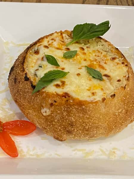This image shows a cheesy baked pasta dish served in a round bread bowl, garnished with fresh basil leaves and accompanied by cherry tomato slices on a white plate. The golden, bubbly cheese topping adds a deliciously crispy finish.