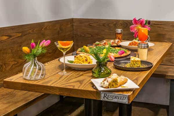 Reserved booth table with pasta, arugula pizza, bruschetta, cannoli, pistachio cake, and cocktails garnished with flowers.
