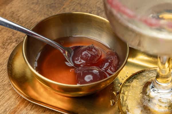 Brass bowl of cocktail cherries in syrup with a small spoon, served alongside a cold drink on a golden tray in a stylish bar setting.